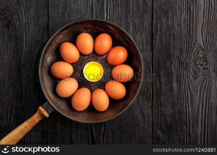 Bright chicken yolk of a broken egg surrounded by chicken eggs in an old cast-iron pan, which is standing on an old black wooden surface, top view, copy space.. Ten brown chicken eggs in an old cast-iron skillet look in the center at a broken egg with a bright yolk.