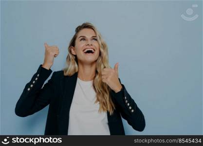 Bright laughing young girl slightly tilted her head back showing thumbs up ok gesture with both hands, happy to approve or to recommend something very good, posing isolated over blue studio wall. Bright laughing young woman showing thumbs up gesture with both hands