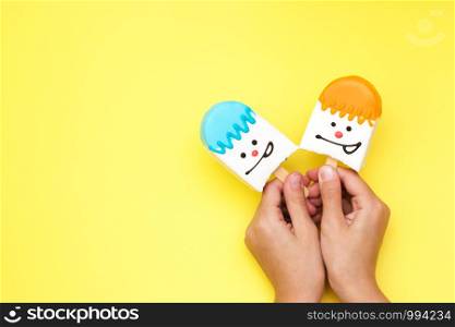 bright multicolored souffle dessert in white chocolate on a stick in hand with a smiley face on a yellow background dessert candies selective focus.. bright multicolored souffle dessert in white chocolate on a stick in hand with a smiley face on a yellow background dessert candies selective focus