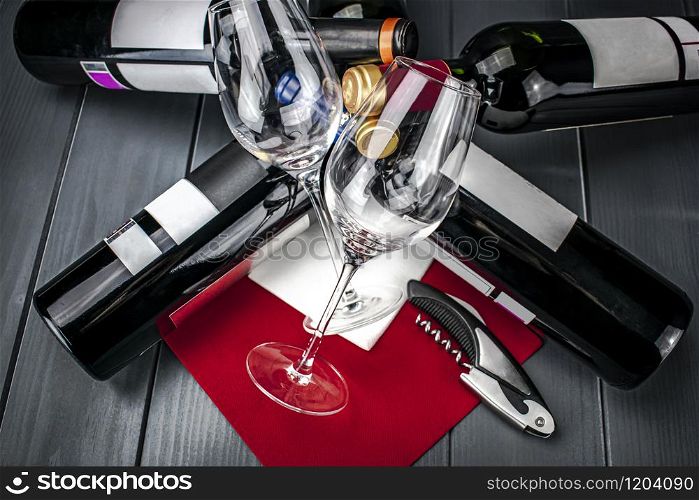 Bright wine glasses lying on bed of bottles of red wine on red tablecloth, corkscrew, napkin and gray wooden background