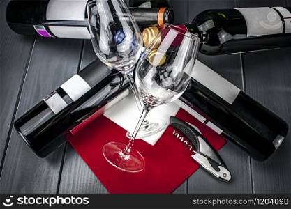 Bright wine glasses lying on bed of bottles of red wine on red tablecloth, corkscrew, napkin and gray wooden background