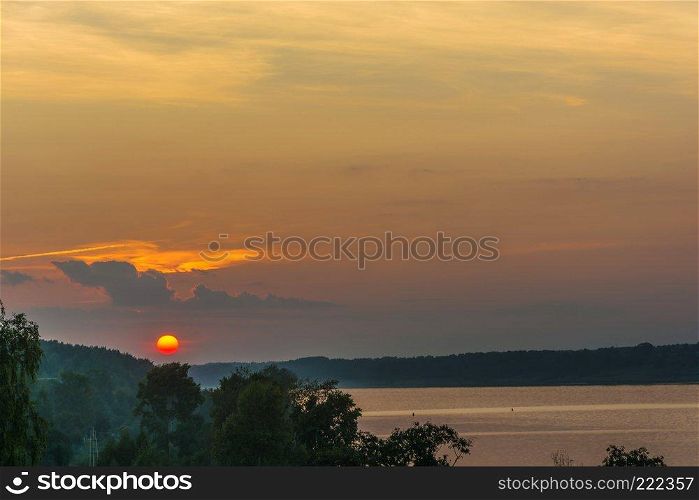 Bright yellow disk of the setting sun against the background of the cloudy sky and green trees on the bank of the Volga River. 