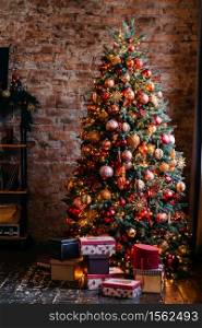 Brightly decorated Christmas tree with bunch of gift boxes near it standing in front of brick wall. . Christmas tree and presents