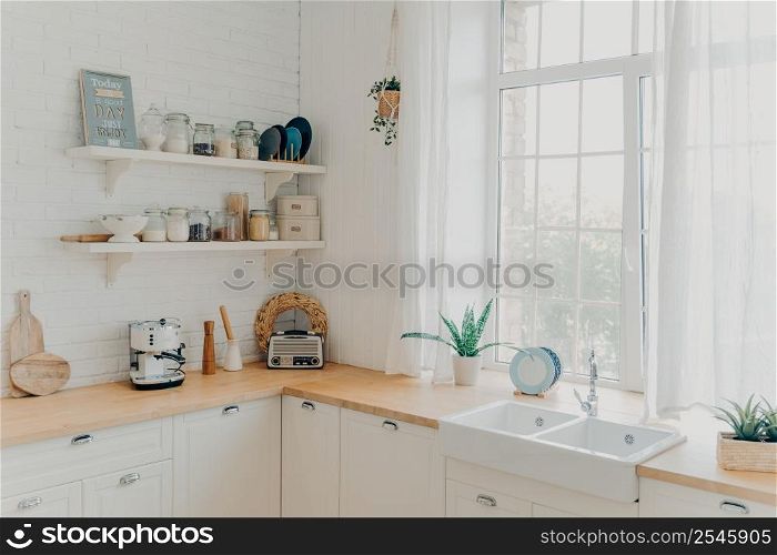 Brightly lit scandinavian kitchen interior. Large window in front of sink. Clean wooden counters along the walls. Accessories and pot plants. Concept of mortgage and moving to modern apartment.. Brightly lit scandinavian kitchen with large window. Concept of mortgage and modern apartment.