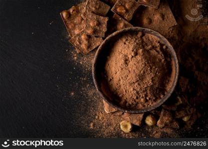 Broken chocolate nuts pieces and cocoa powder in bowl on dark stone background