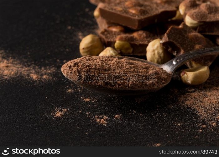 Broken chocolate nuts pieces and cocoa powder in spoon on dark background