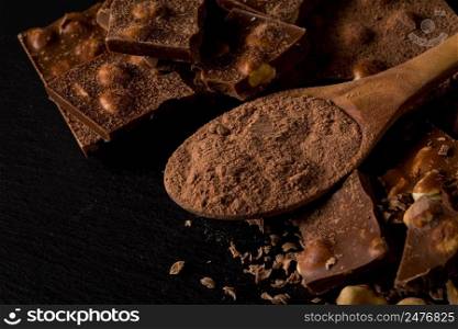 Broken chocolate nuts pieces and cocoa powder in spoon on dark stone background
