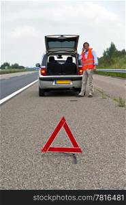 Broken down car at the side of the road, with a man, wearing a reflective vest calling for assistance, a warning triangle. proper procedrue of road safety