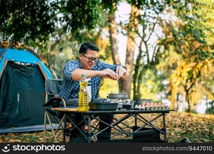 Broken egg falls into the frying pan at the c&site. Happy Asian traveler man cooking with tent background. Outdoor cooking, traveling, c&ing, lifestyle concept.