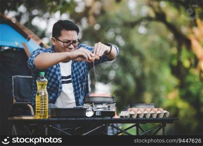Broken egg falls into the frying pan at the campsite. Happy Asian traveler man cooking with tent background. Outdoor cooking, traveling, camping, lifestyle concept.