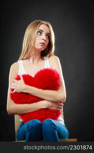 Broken heart love concept. Sad unhappy woman sitting on chair hugging red heart pillow dark background