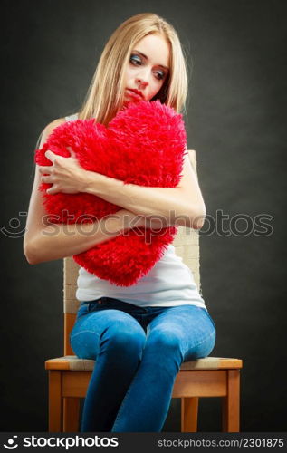 Broken heart love concept. Sad unhappy woman sitting on chair hugging red heart pillow dark background