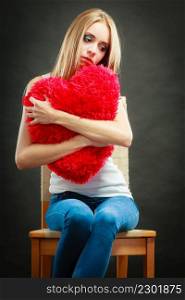 Broken heart love concept. Sad unhappy woman sitting on chair hugging red heart pillow dark background