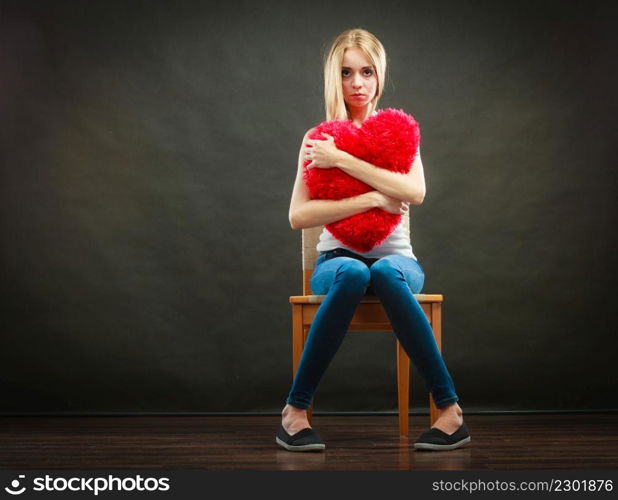 Broken heart love concept. Sad unhappy woman sitting on chair hugging red heart pillow dark background