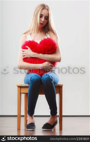 Broken heart love concept. Sad unhappy woman sitting on chair hugging red heart pillow