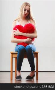 Broken heart love concept. Sad unhappy woman sitting on chair hugging red heart pillow