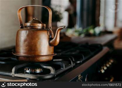 Bronze kettle in modern kitchen. Old vintage teapot on gas stove. Preparing tea. Aluminium teakettle. Sunny daylight from window.