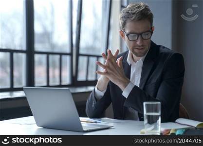 Brooding concentrated young German businessman in formal suit and glasses looks through notes from agenda last time on his desk, focused male worker working on laptop online. Computer work concept. Brooding concentrated German businessman in formal suit and glasses sitting in office