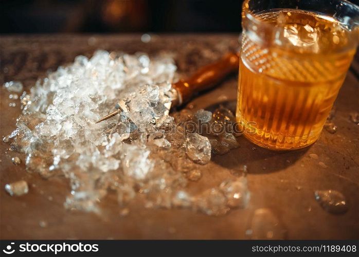 Brown beverage in a glass and ice on bar counter closeup, nobody. Refreshing of alcoholic drink, cold crystals on the table. Brown beverage in a glass and ice on bar counter