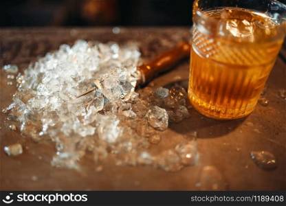 Brown beverage in a glass and ice on bar counter closeup, nobody. Refreshing of alcoholic drink, cold crystals on the table. Brown beverage in a glass and ice on bar counter