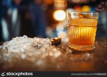 Brown beverage in a glass and ice on bar counter closeup, nobody. Refreshing of alcoholic drink, cold crystals on the table. Brown beverage in a glass and ice on bar counter