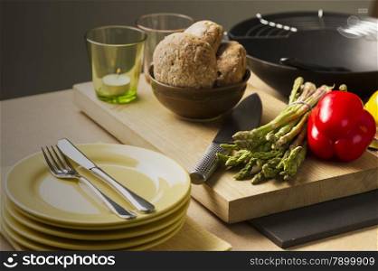 Brown Bread on Bowl, Fresh Veggies, Cutting Knife, Glass with Candles and Plates with Fork and Knife on top of Table. Preparing for Meal.