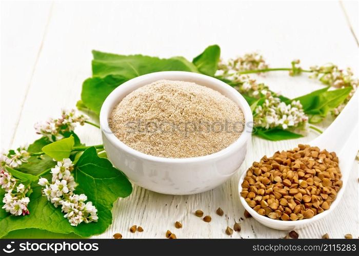 Brown buckwheat flour in a bowl, brown groats in a spoon, flowers and buckwheat leaves on wooden board background