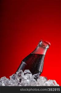 Brown carbonated drink in a wet bottle with a red cap in ice cubes on a red background. Brown carbonated drink in wet bottle with red cap in ice cubes on red background