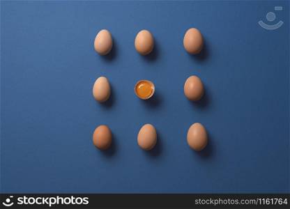 Brown eggs and one cracked egg with yolk, aligned on a blue paper background. Flat lay of organic eggs in symmetry. One-half egg and many whole eggs.