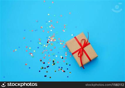 brown gift box wrapped in paper and tied with silk ribbon on a blue background with multi-colored shiny confetti, festive backdrop for birthday, valentine&rsquo;s day