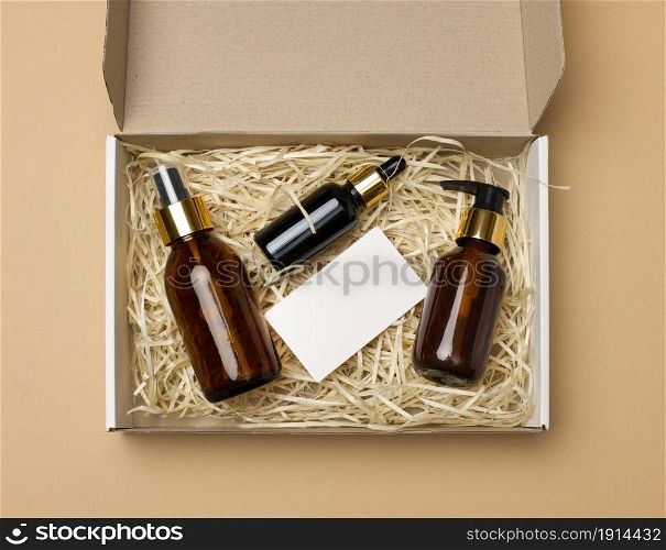 brown glass bottles for cosmetics and a white paper blank business card lie in a cardboard paper box on a beige background, top view. Delivery