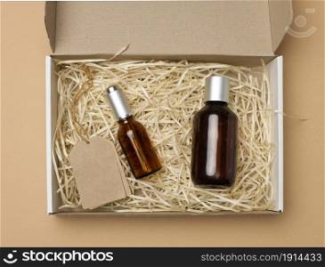brown glass bottles for cosmetics and paper tags on a rope lie in a cardboard paper box on a beige background, top view
