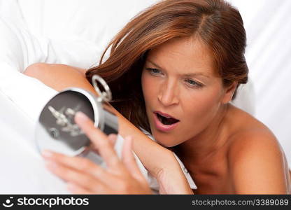 Brown hair woman yawning and holding alarm clock in white bed