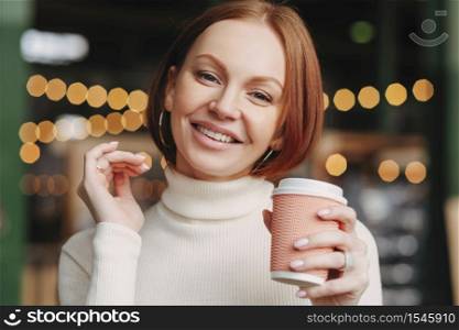 Brown haired woman with make up, toothy smile, dressed in white turtle neck sweater, enjoys hot drink, smiles positively, poses in coffee shop, has make up, expresses happiness. Drinking concept