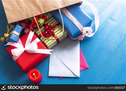 Brown paper bag overturned on a blue wooden table, while multicolored presents and closed envelopes come out of it, near a red, lit candle.