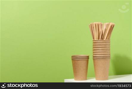 brown paper cardboard cups and wooden stirring sticks on a white table, green background. Eco-friendly tableware, zero waste