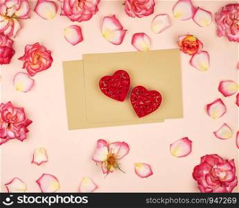 brown paper envelopes and two red carved hearts in the middle of rosebuds, top view, festive backdrop