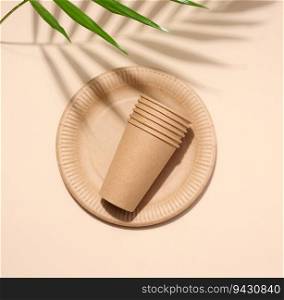 Brown paper plate, stack of paper cups, disposable tableware on a beige background, top view