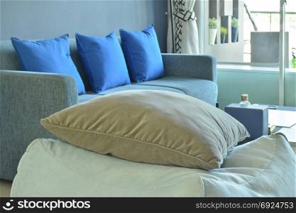 Brown pillow on beige fabric cubic stool with blue sofa in background