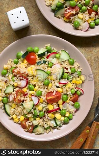 Brown rice salad with cherry tomato, corn, cucumber, radish and peas served on plate, photographed overhead on slate with natural light