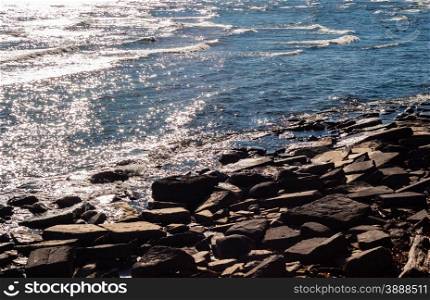 Brown rocks and stones diagonally at water?s edge, with small waves breaking and water reflecting bright sunlight.