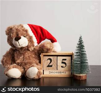 brown teddy bear in red hat, desk wooden calendar with the date december 25 and green decorative tree, christmas background