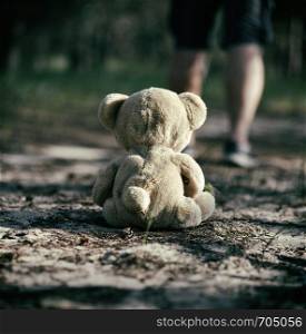 brown teddy bear sits back in the middle of a sandy road in the forest, concept of loneliness and forgetting