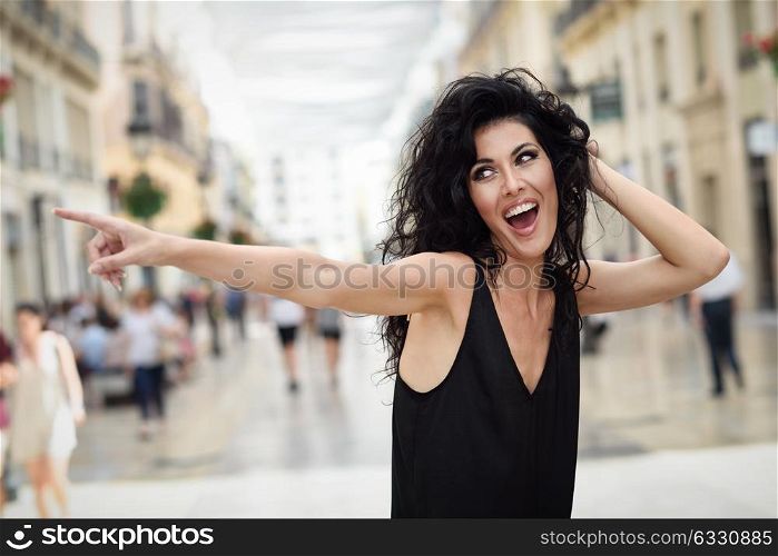 Brunette funny woman wearing casual clothes pointing with her finger in the street. Young girl with curly hairstyle standing in urban background