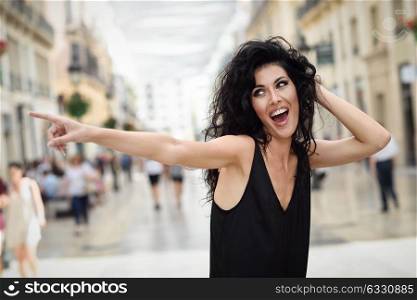 Brunette funny woman wearing casual clothes pointing with her finger in the street. Young girl with curly hairstyle standing in urban background