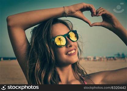 Brunette girl on beach sunglasses with palm tree reflection and fingers heart shape gesture
