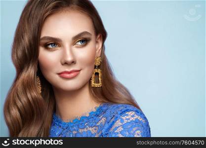 Brunette girl with perfect makeup. Beautiful model woman with curly hairstyle. Care and beauty hair products. Lady with fashionable gold makeup. Model with jewelry on blue background