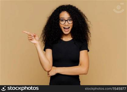 Brunette mixed race woman points aside with index finger, has glad curious expression, promots awesome product, dressed in casual clothes, isolated over brown background. People and advertisement