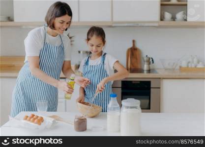 Brunette mother adds oil in dough, little daughter helps to make pastry, whisks ingredients, pose together against kitchen interior, prepare bakery together. Small helper with mommy. Homemade food
