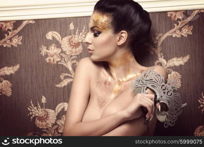 brunette sexy woman covering her naked breast and posing with glossy golden make-up and glitter carnival mask in the hand, creative hairdo. Turned on her profile